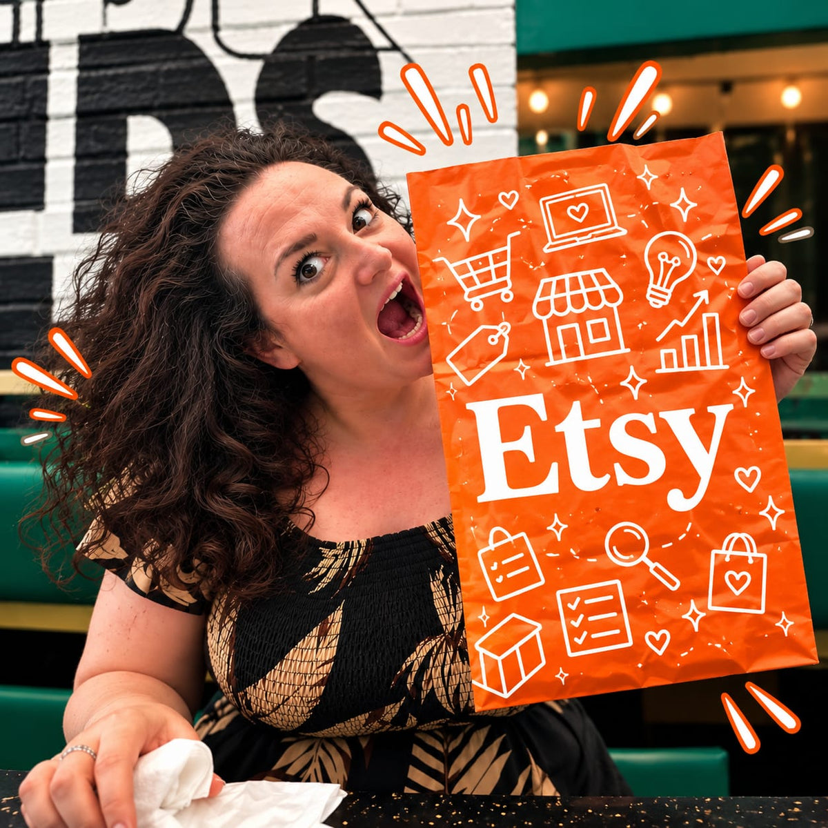 A woman with curly dark hair holds up an orange paper shopping bag printed with white Etsy icons and the word 'Etsy' in large white letters. She is smiling and looking toward the camera in what appears to be a restaurant or cafe setting with green seating and a black-and-white wall behind her.